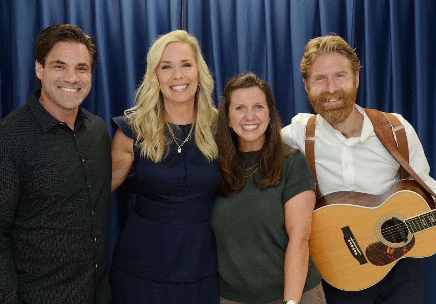 Four people posing together in front of a blue curtain, with an acoustic guitar